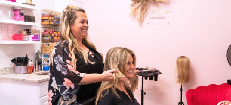 Hairstylist styling a smiling client’s long blonde hair in her salon suite